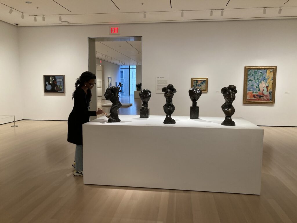 Efeh holds a large brush and dusts the base of a sleek glossy black bust sculpture on a pedestal in an art gallery. Four other glossy black bust sculptures fill the pedestal.