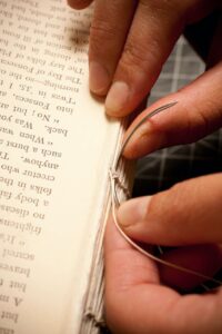 Hands hold a threaded curved needle next to a sewn binding for a book