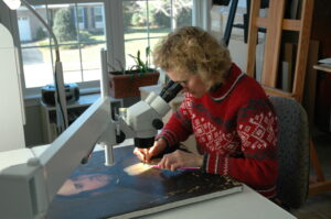 Nancy sits at a microscope positioned over a painting paid horizontally on a worksurface.