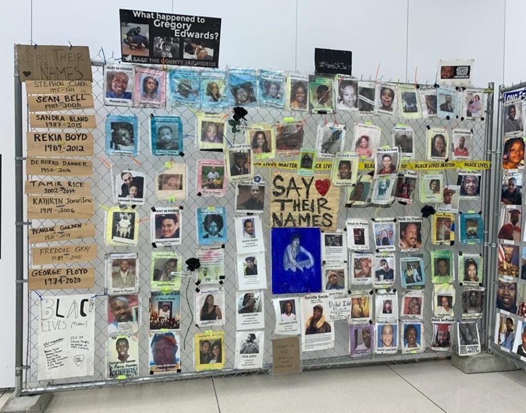 Image of silver chain-link fence with attached memorial signs and protest artifacts that comprised the Black Lives Matter Memorial Fence installation.