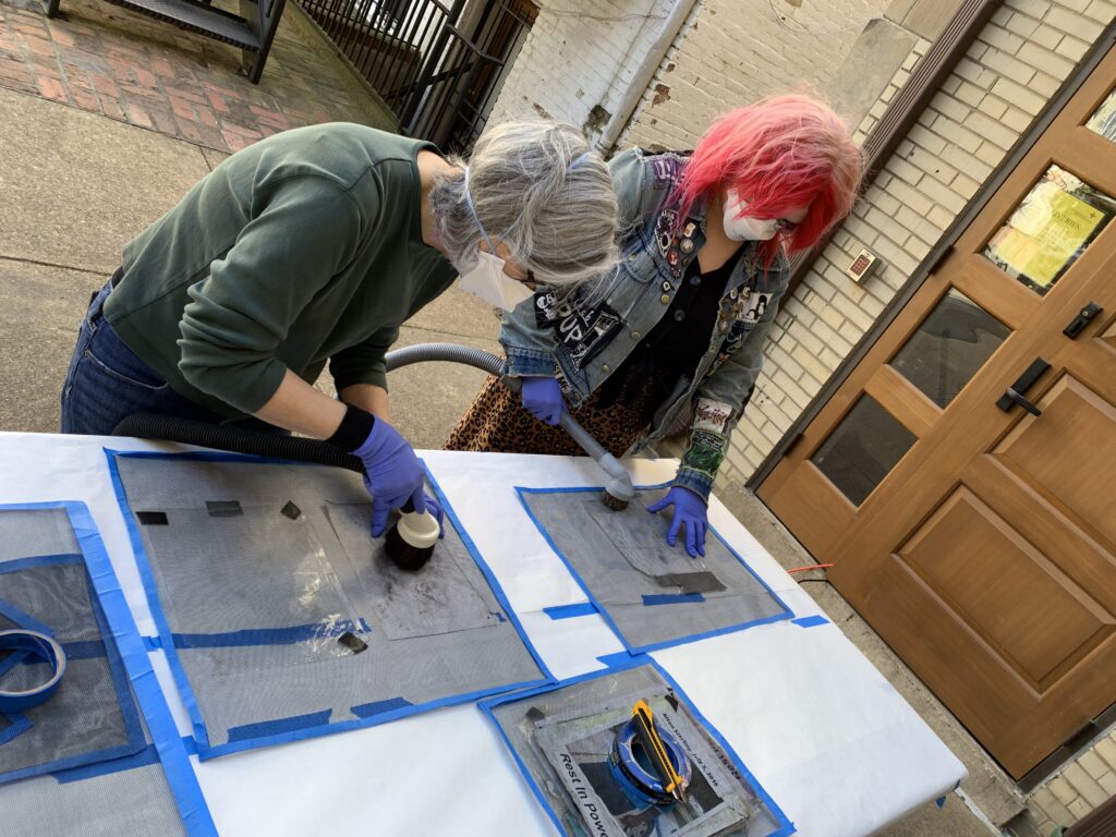 Two people bending over a table working to brush vacuum triage objects through a protective screen