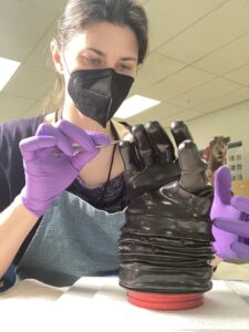 A professional using a pair of tweezers to clean the rubber surfaces of a black spacesuit glove, which is standing up on a workbench.