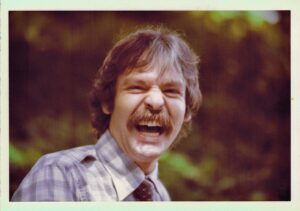 A man photographed outside smiling widely with a moustache and plaid shirt.