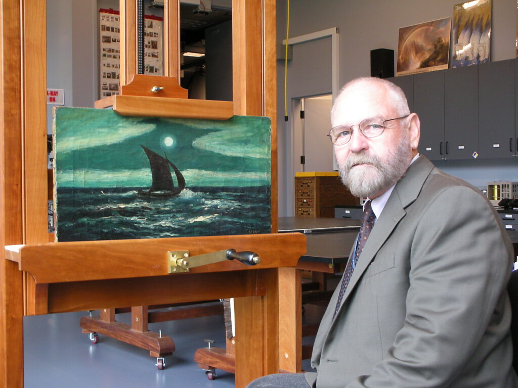 A conservator sitting beside a painting of a boat on water, primarily blue-green color scheme. The conservator is in a gray suit and glasses and looks over their shoulder at the camera.