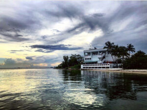 Wide vista of a shoreline with purple, blue stormclouds over a sunset, above a structure on the shore with palm trees.