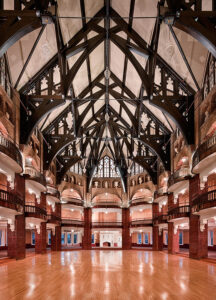 A large, vaulted hallway with multiple floors with dark wood forming a truss-and-tie-beam roof supported by red-brick columns.